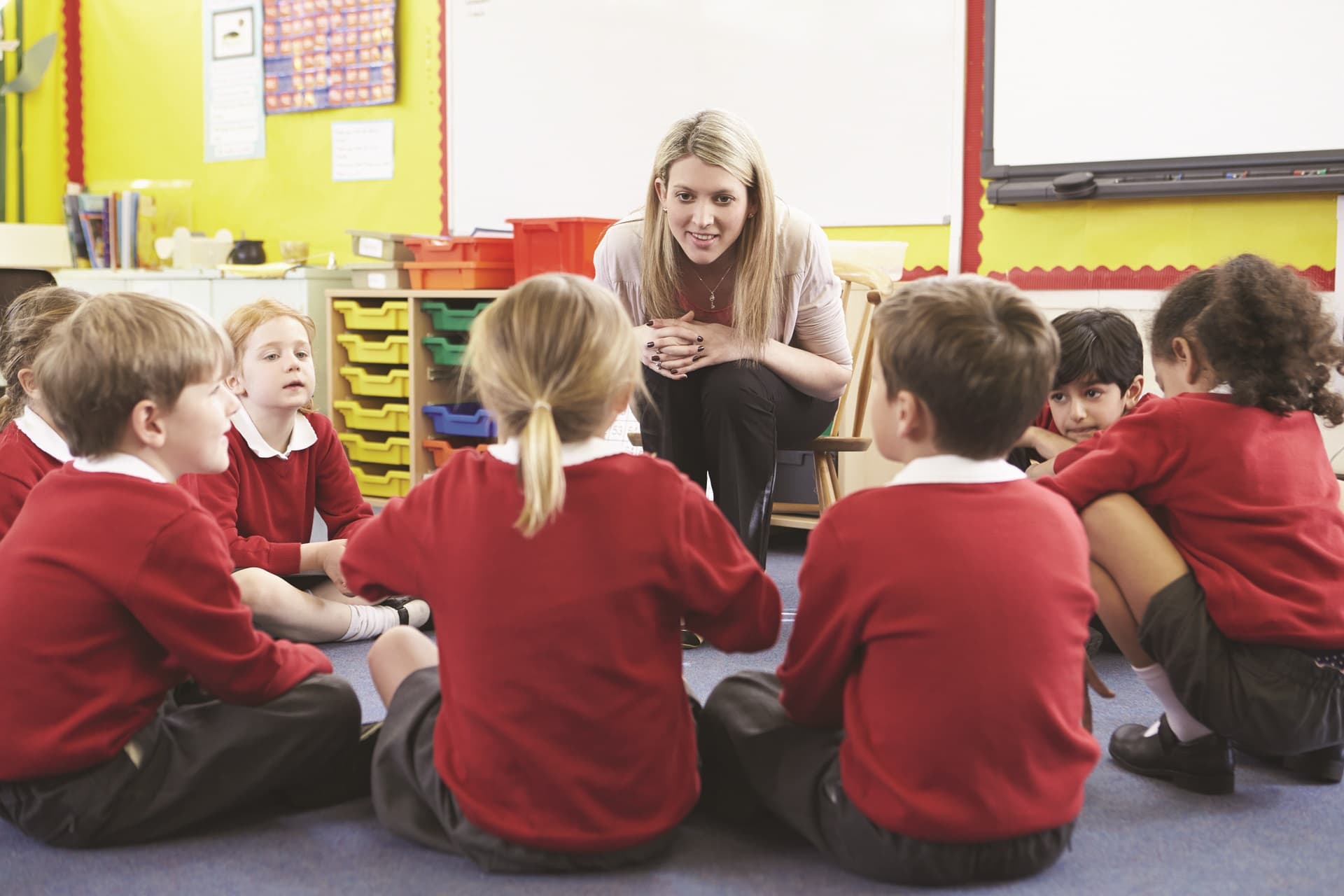 Image of a classroom with teachers and students. The teacher is actively working with a student, while other students are participating in the activity. The classroom is well-lit with posters on the walls.
