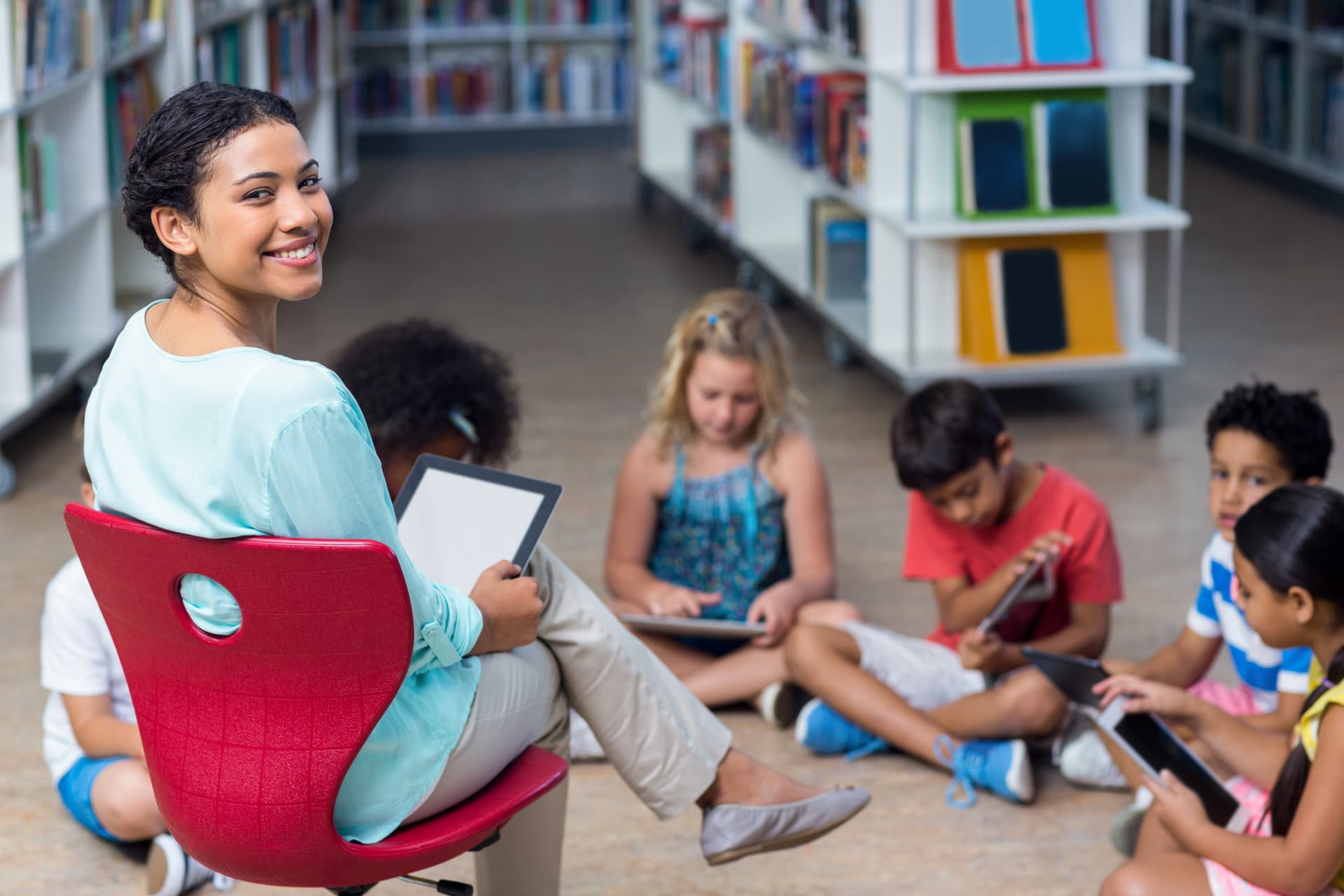 Teacher with tablet supervising diverse students using digital devices in school library